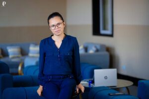 Une femme, vêtue d'un ensemble bleu foncé, est assise sur un canapé dans un espace de bureau moderne et lumineux. Elle porte des lunettes et regarde l'objectif.